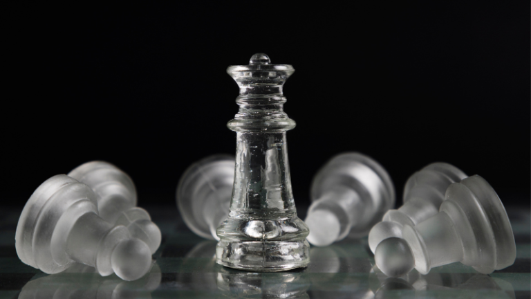 Transparent glass chess king standing upright while other chess pieces lie fallen on a reflective surface against a dark background.
