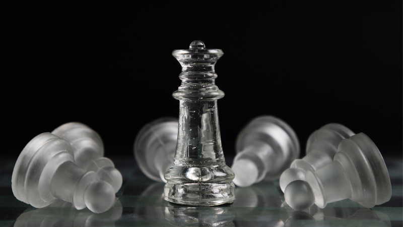 Transparent glass chess king standing upright while other chess pieces lie fallen on a reflective surface against a dark background.