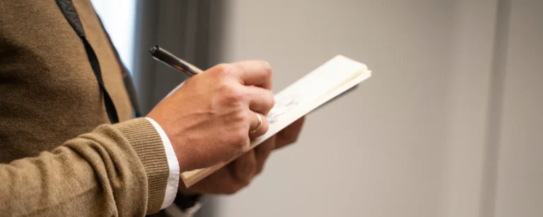 Close-up of a person in a beige sweater and white shirt writing notes with a pen in a small notebook while standing indoors.