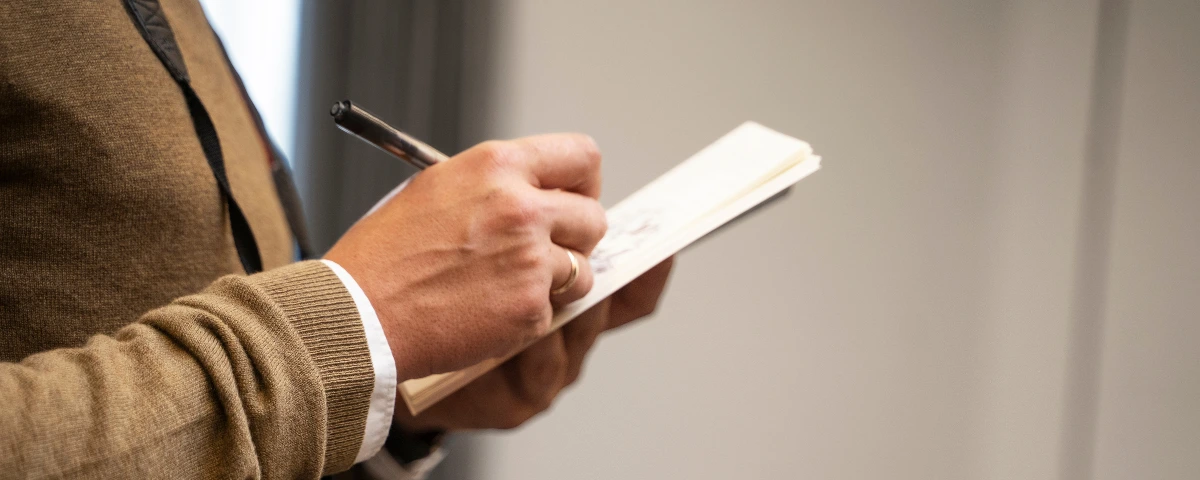 Close-up of a person in a beige sweater and white shirt writing notes with a pen in a small notebook while standing indoors.