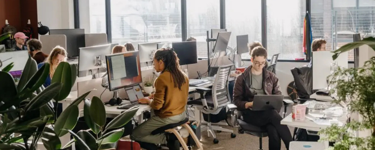 Employees work at computer stations in an open-plan office with large windows and collaborative workspaces.
