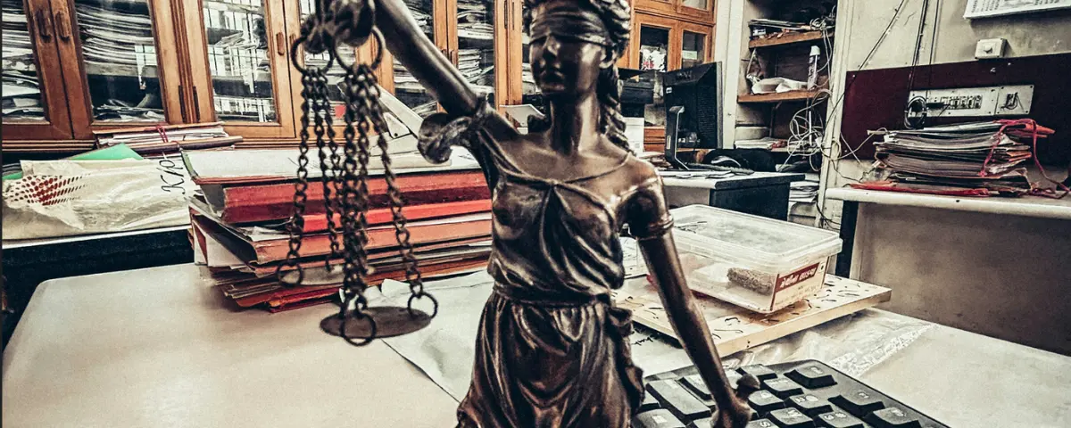 Blindfolded Lady Justice statue holding scales on a desk surrounded by stacks of bound case files and a computer keyboard in an Indian court office, illustrating AI in Indian courts