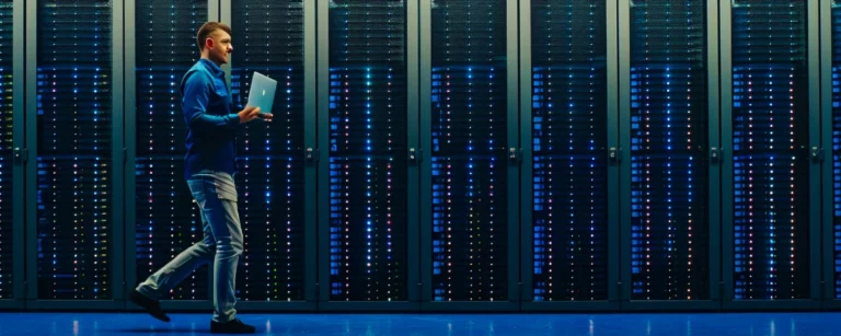 IT technician carrying a laptop walks through a brightly lit modern data centre filled with rows of illuminated server rackS.