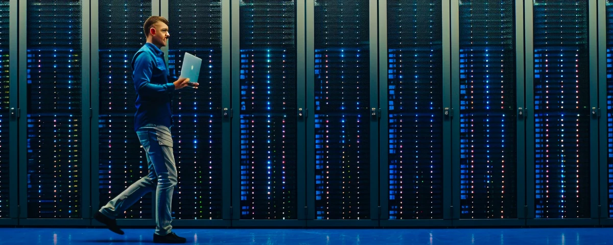 IT technician carrying a laptop walks through a brightly lit modern data centre filled with rows of illuminated server rackS.