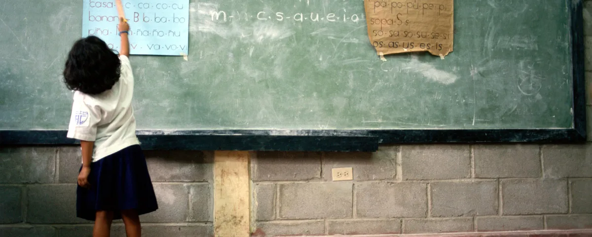 Girl in school uniform points to a syllable chart on a classroom blackboard, illustrating the World Bank AI report