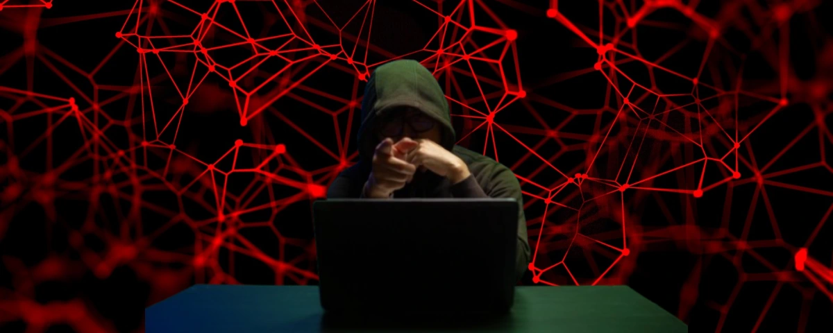 Hooded individual using a laptop against a red digital network illustrating AI-assisted cyberattacks and data theft.